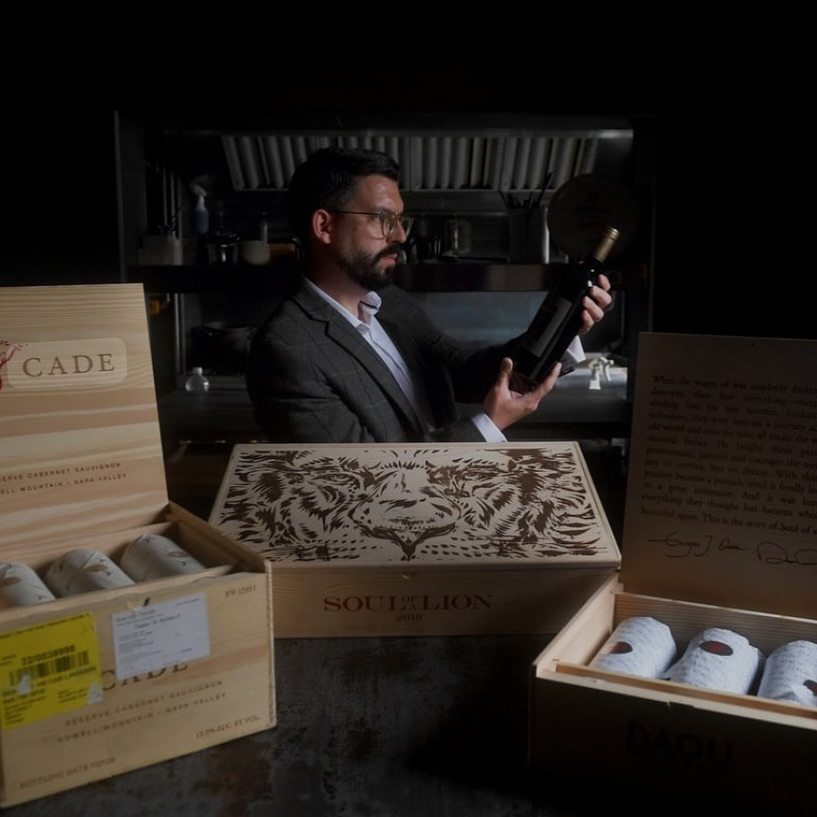 Person holding a wine bottle in a dimly lit setting, surrounded by open wooden crates containing wrapped bottles. 