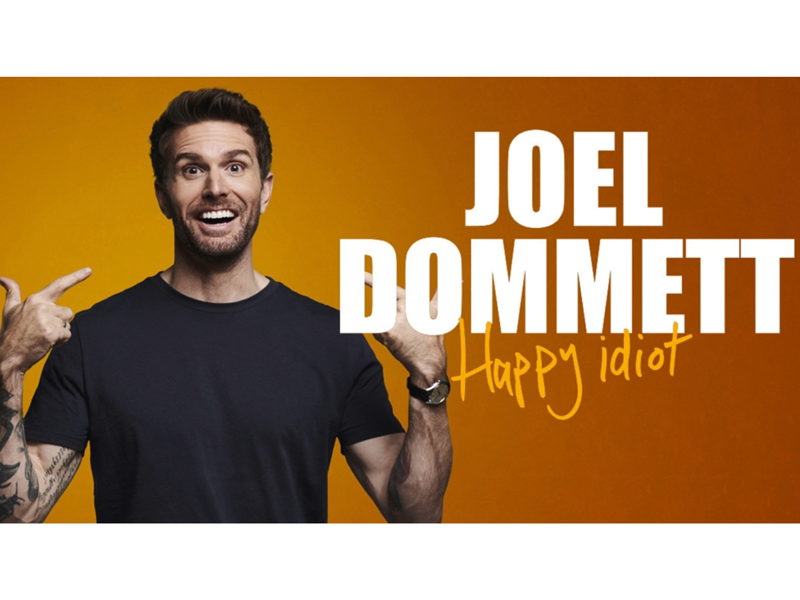 Joel Dommett is smiling and pointing at himself. Next to him it says 'Joel Dommett - Happy Idiot'.