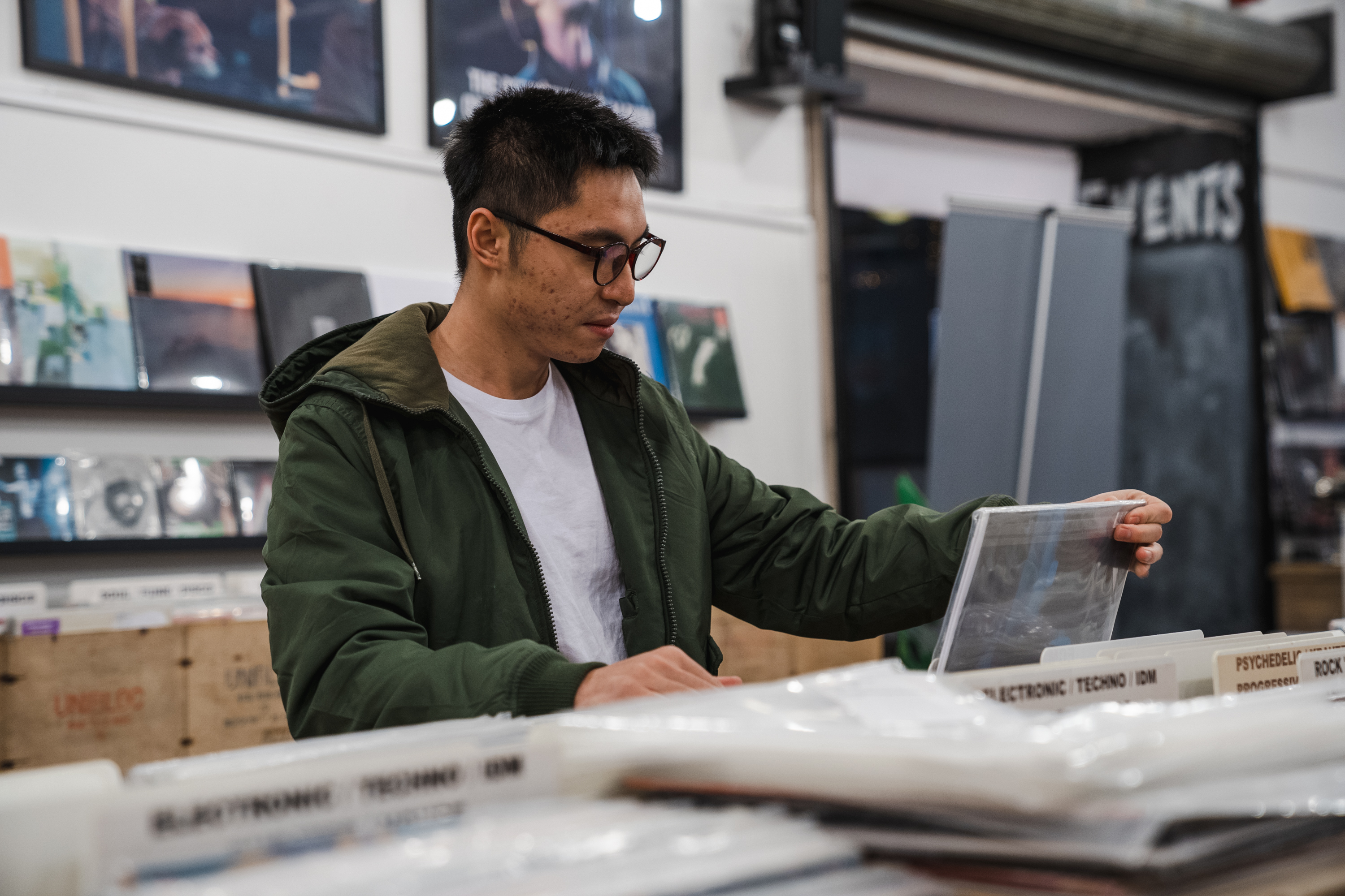 A person wearing a white T-shirt and a green jacket is browsing through vinyl records in a record store. The individual is holding one record while looking through a bin labeled with music genres such as "Electronic Techno" and "Psychedelic Rock." The background shows shelves with more records and framed artwork on the wall.