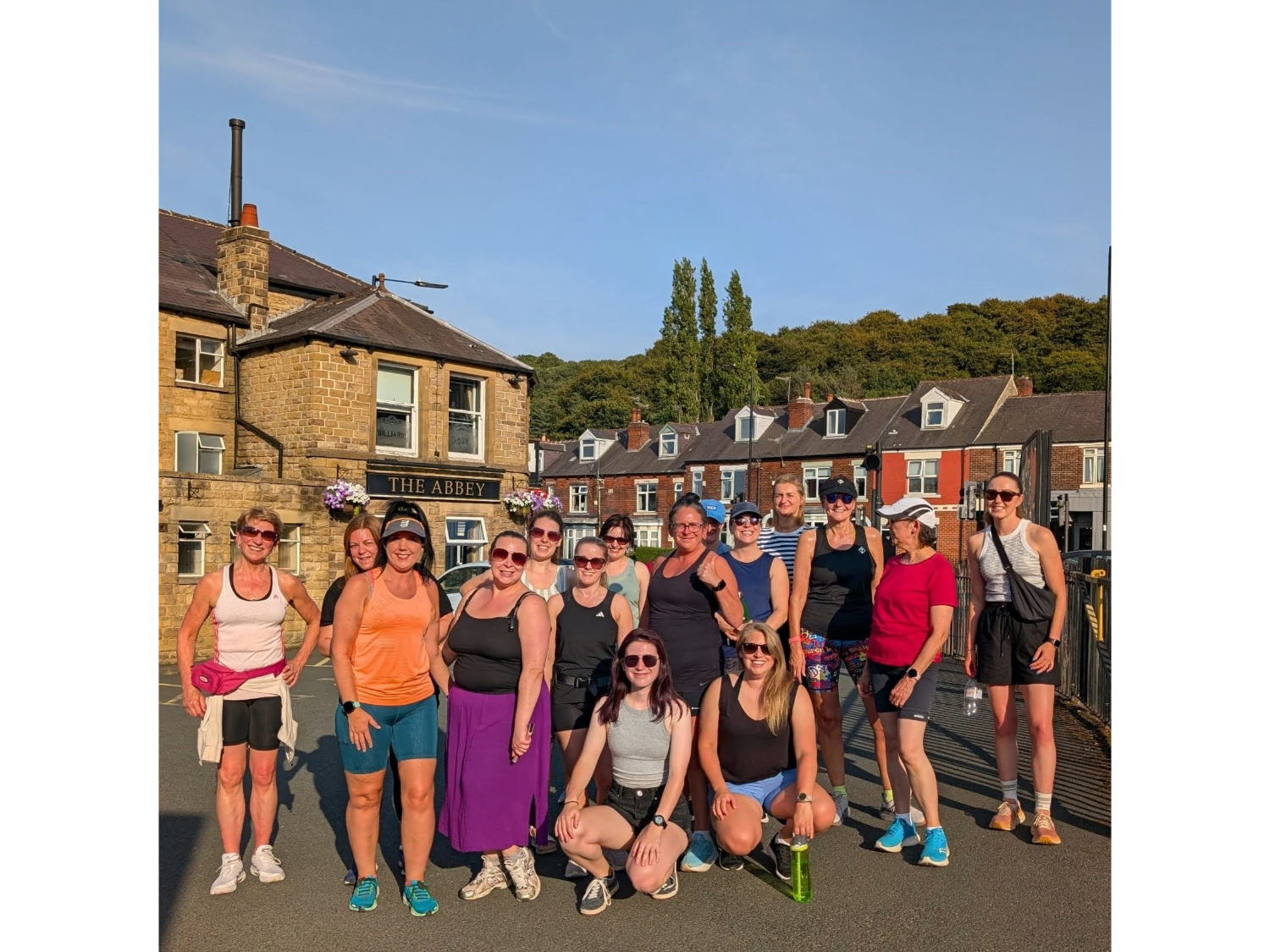 Group of 17 people in casual athletic wear standing on a street in front of a building named "The Abbey." The background includes houses and greenery on a hillside under sunny, clear weather.