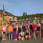 Group of 17 people in casual athletic wear standing on a street in front of a building named "The Abbey." The background includes houses and greenery on a hillside under sunny, clear weather.