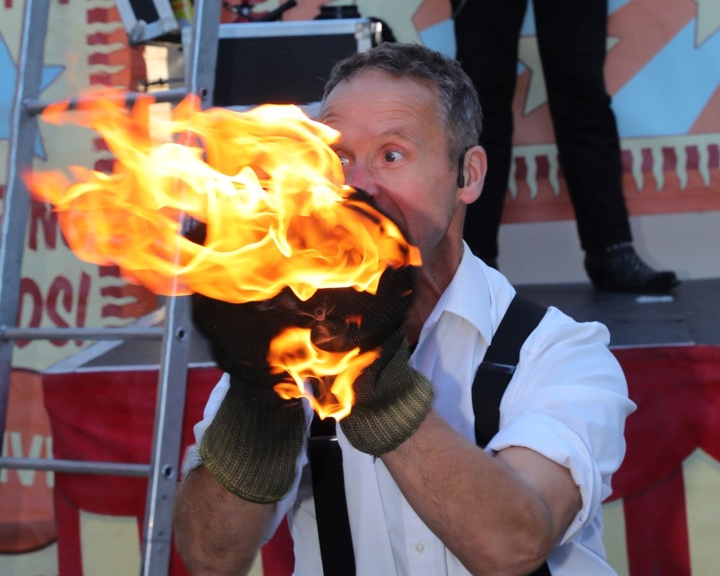 A man dressed in a white shirt and braces breathes fire.