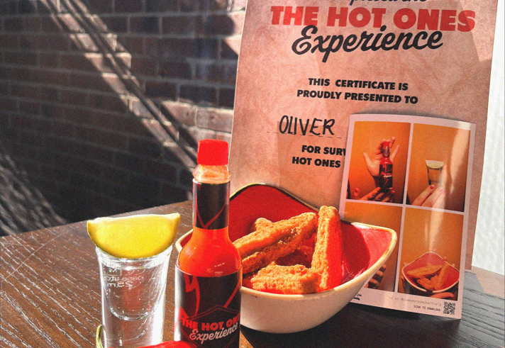 A table with a menu, a shot of spirits, a bottle of hot sauce and some red chillies. 