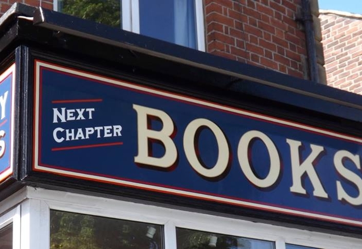 Close-up view of the Next Chapter Books shop sign, featuring bold cream lettering on a dark blue background with red and white borders. The phrases ‘WE BUY BOOKS’ and ‘NEXT CHAPTER BOOKS’ are displayed on the corner fascia of a red‑brick building.
