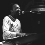 A black and white photo of Jalen Ngonda sat at a piano, playing and singing.