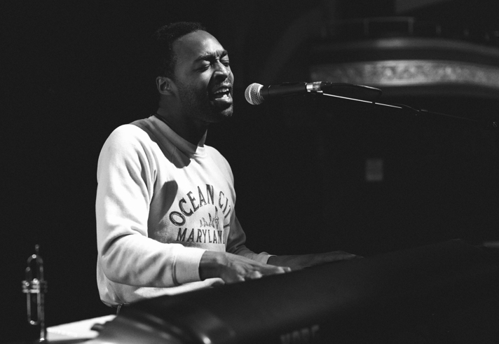 A black and white photo of Jalen Ngonda sat at a piano, playing and singing.