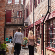 Two people walking along a cobbled courtyard lined with red‑brick buildings and small shopfronts, with potted plants, windows and doors creating a sheltered, historic city‑centre passageway.