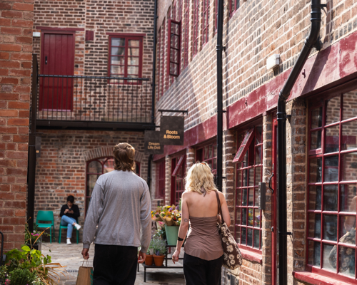 Two people walking along a cobbled courtyard lined with red‑brick buildings and small shopfronts, with potted plants, windows and doors creating a sheltered, historic city‑centre passageway.
