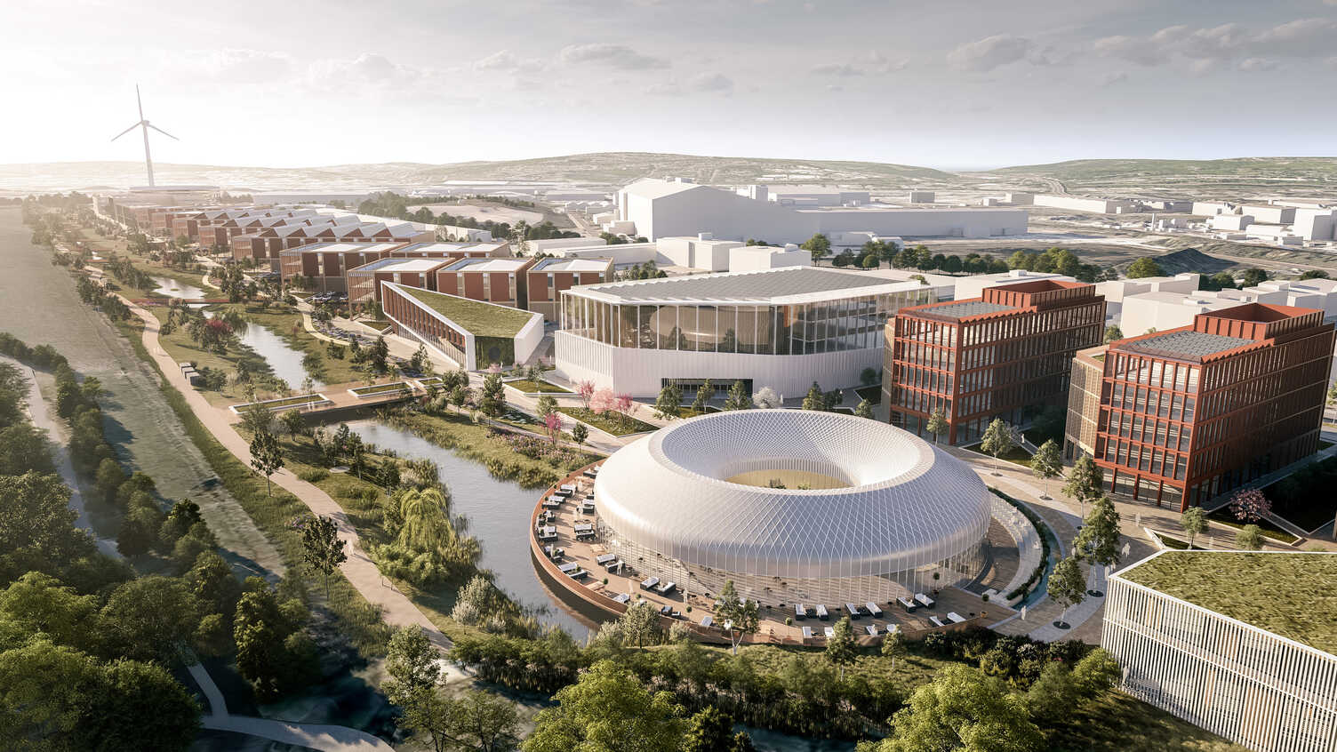 “Aerial view of a modern urban development featuring a circular glass building surrounded by water, red-brick office blocks, and landscaped green spaces. A canal runs through the area, with rows of residential buildings on the left and a large wind turbine visible in the distance.”