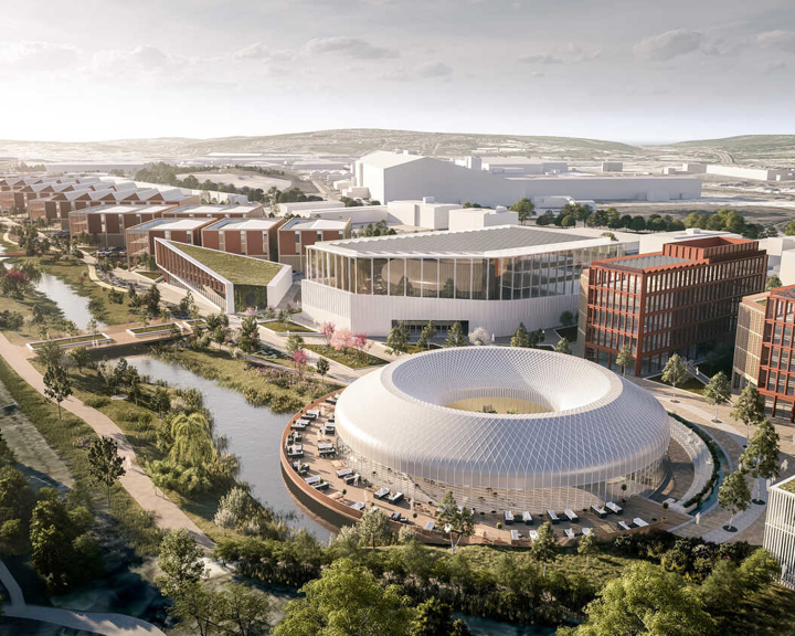 “Aerial view of a modern urban development featuring a circular glass building surrounded by water, red-brick office blocks, and landscaped green spaces. A canal runs through the area, with rows of residential buildings on the left and a large wind turbine visible in the distance.”