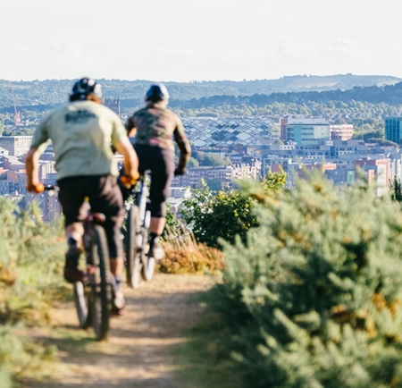 Two cyclists riding mountain bikes along a dirt trail surrounded by green shrubs. The path leads toward a scenic view of a city in the background, with buildings, cranes, and rolling hills visible under a clear sky. The image captures an outdoor adventure with an urban landscape in the distance.