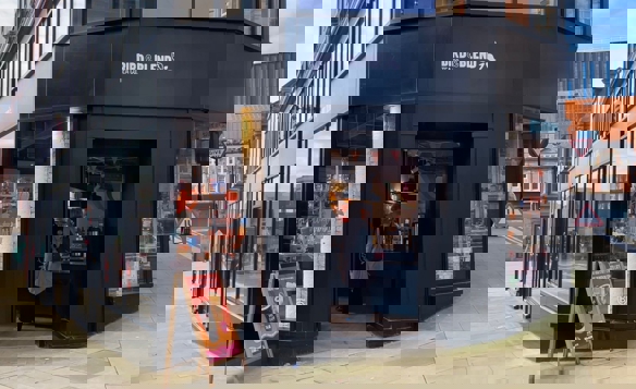 Exterior view of a corner tea shop called “Bird & Blend Tea Co.” with large glass windows and a black storefront. The entrance is open, revealing a colorful interior with hanging decorations and a counter. A sandwich board outside displays promotional drinks, and the shop is located on a paved street with buildings and traffic signs visible in the background.