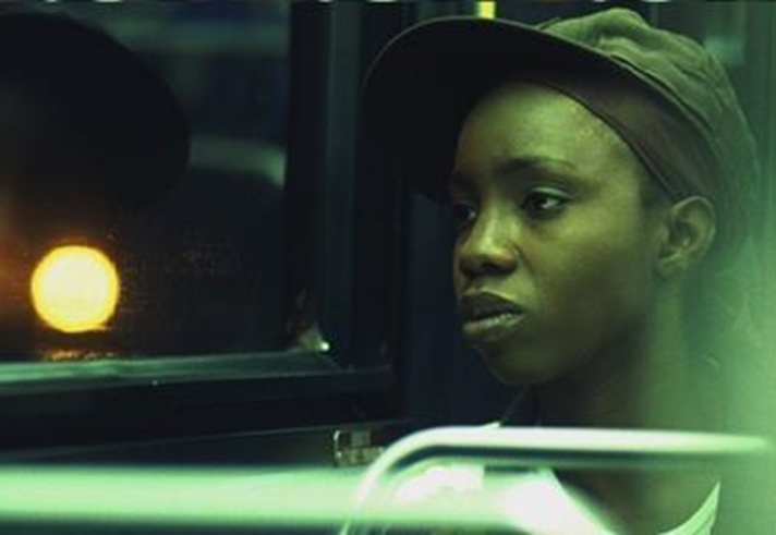 A person wearing a hat sits on a bus, gazing out of the window. Their reflection is visible in the glass, illuminated by a bright light outside, creating a glowing effect. The scene evokes a quiet moment of contemplation.