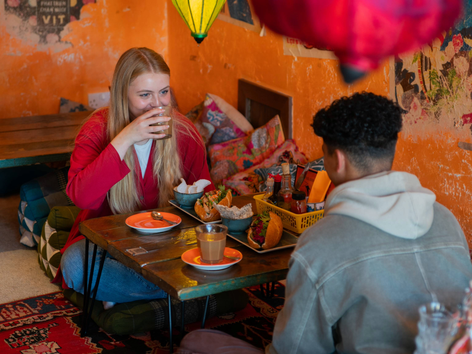 Two people are seated on cushions on the floor, at a low table, enjoying the meal. There are brightly coloured lanterns hanging from the ceiling. The orange walls are decorated with old and faded posters.