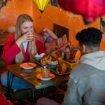 Two people are seated on cushions on the floor, at a low table, enjoying the meal. There are brightly coloured lanterns hanging from the ceiling. The orange walls are decorated with old and faded posters.