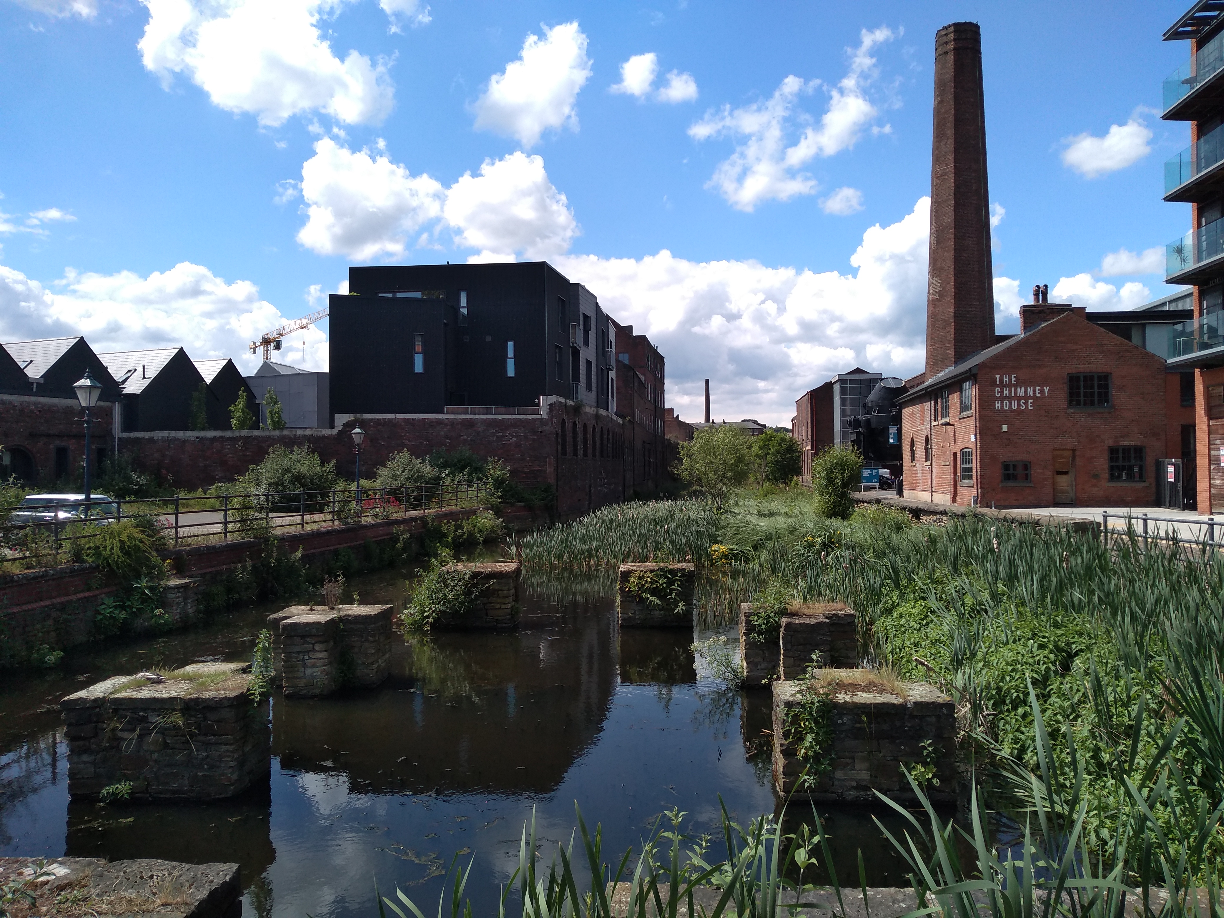 Goit With Chimney House Little Kelham - View of a canal with stone foundations and tall green reeds in the foreground, surrounded by brick buildings and modern apartments. A tall industrial chimney and a building labeled ‘The Chimney House’ are visible on the right, under a bright blue sky with scattered clouds.