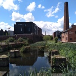 Goit With Chimney House Little Kelham - View of a canal with stone foundations and tall green reeds in the foreground, surrounded by brick buildings and modern apartments. A tall industrial chimney and a building labeled ‘The Chimney House’ are visible on the right, under a bright blue sky with scattered clouds.