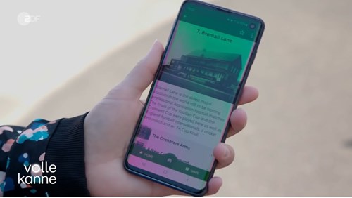 Close-up of a smartphone held in one hand, displaying an app screen with information about ‘7. Bramall Lane’ and a photo of a building. The text mentions The Cricketers Arms, and the phone interface shows navigation icons at the bottom. Text on the bottom left reads ‘volle kanne.’
