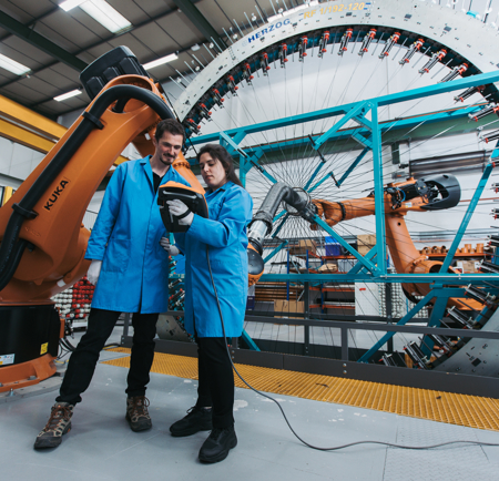 Two people in blue lab coats are talking and looking at a tablet like device. Behind them is an industrial weaving loom.