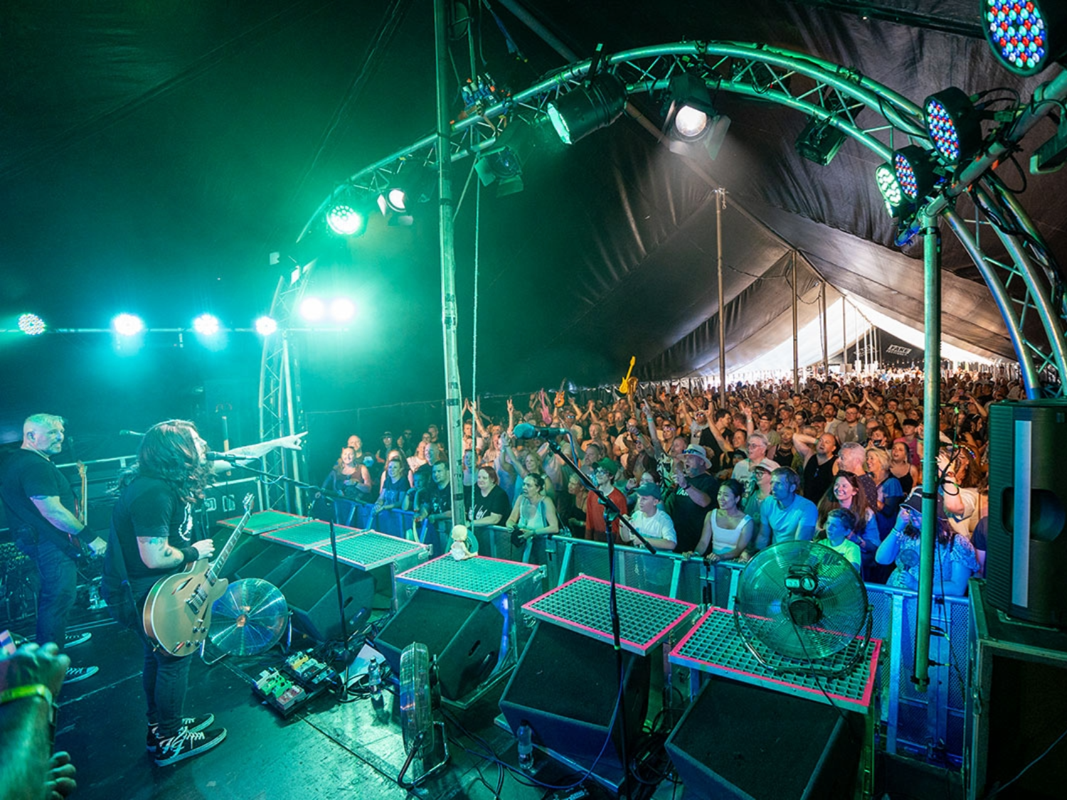 A live band performs under a large tent with bright green stage lights illuminating the musicians and equipment. The guitarist stands near the front, pointing toward a packed audience behind a barrier. The crowd is cheering with raised hands, and the stage setup includes speakers, fans, and lighting rigs overhead.