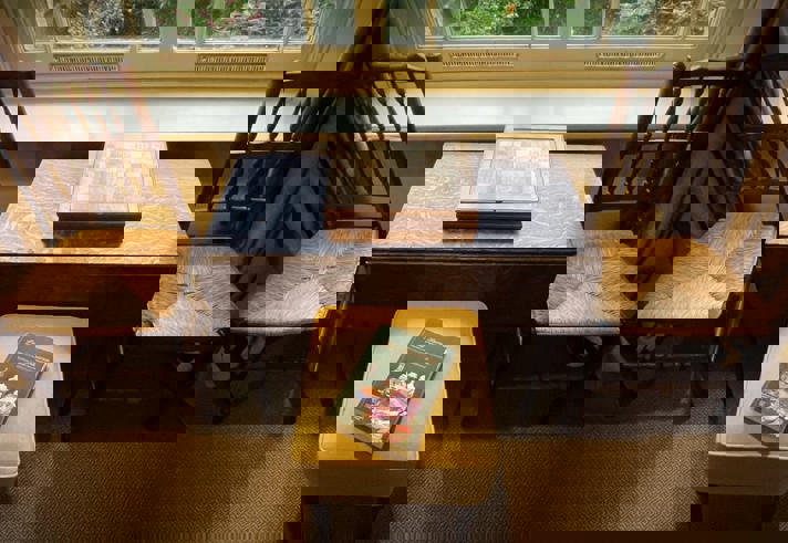 A table with two chairs and a stool, next to a window. There is a chess board on the table and a hard back book on the stool.