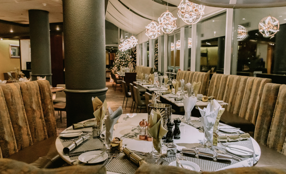 Elegant restaurant interior with round tables set for fine dining, featuring neatly arranged plates, cutlery, folded napkins, and glassware. Plush curved booth seating surrounds the tables, and modern spherical pendant lights hang from the ceiling. Large windows line one side of the room, with a decorated Christmas tree visible in the background, creating a warm and festive atmosphere.