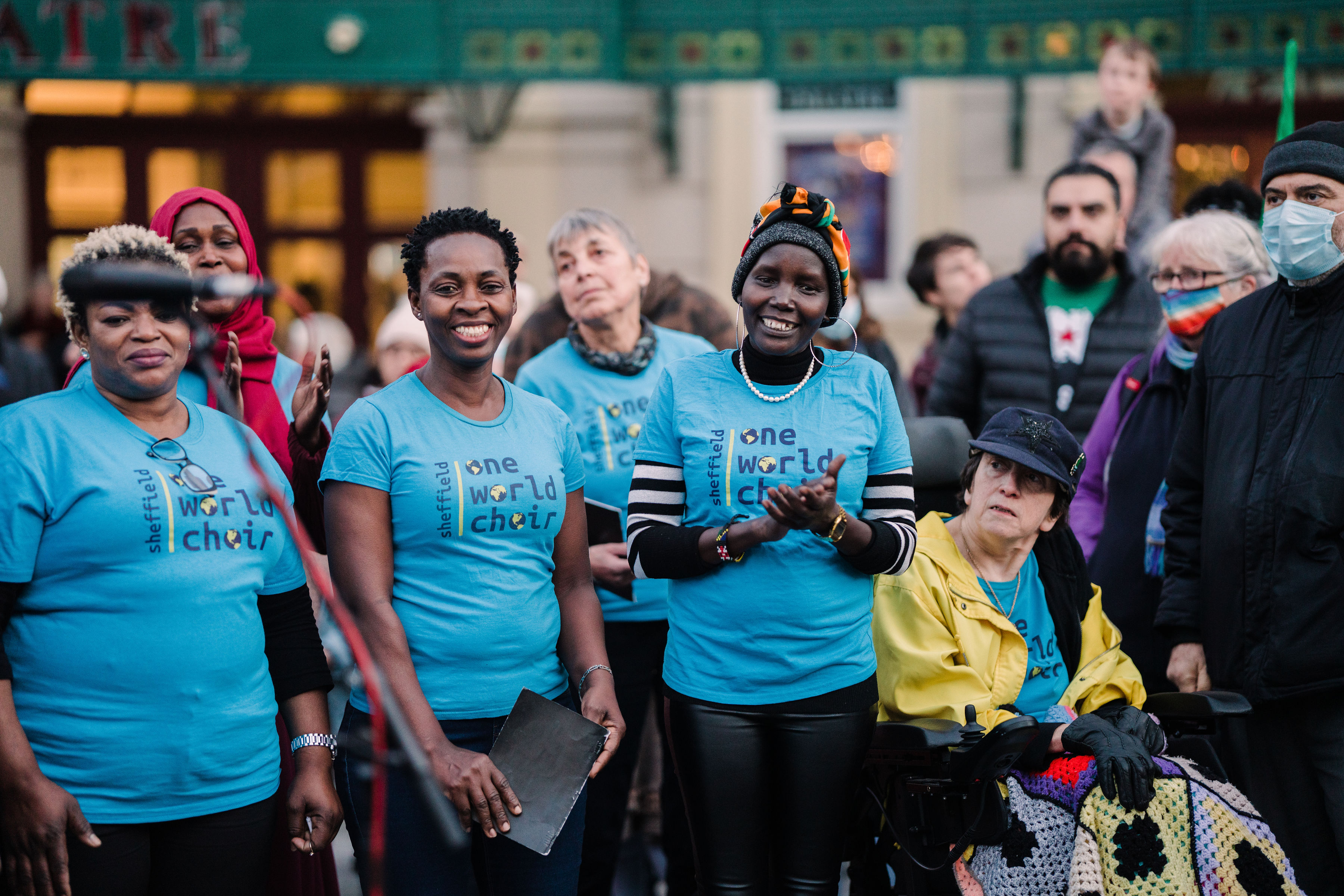 A group of people wearing matching light blue T-shirts with the text “Sheffield One World Choir” printed on them, standing together at an outdoor event. Some individuals are holding items, and one person is seated in a wheelchair with a colorful crocheted blanket. The background shows a building with signage and other attendees gathered around.