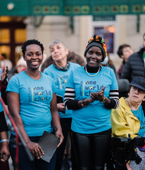 A group of people wearing matching light blue T-shirts with the text “Sheffield One World Choir” printed on them, standing together at an outdoor event. Some individuals are holding items, and one person is seated in a wheelchair with a colorful crocheted blanket. The background shows a building with signage and other attendees gathered around.