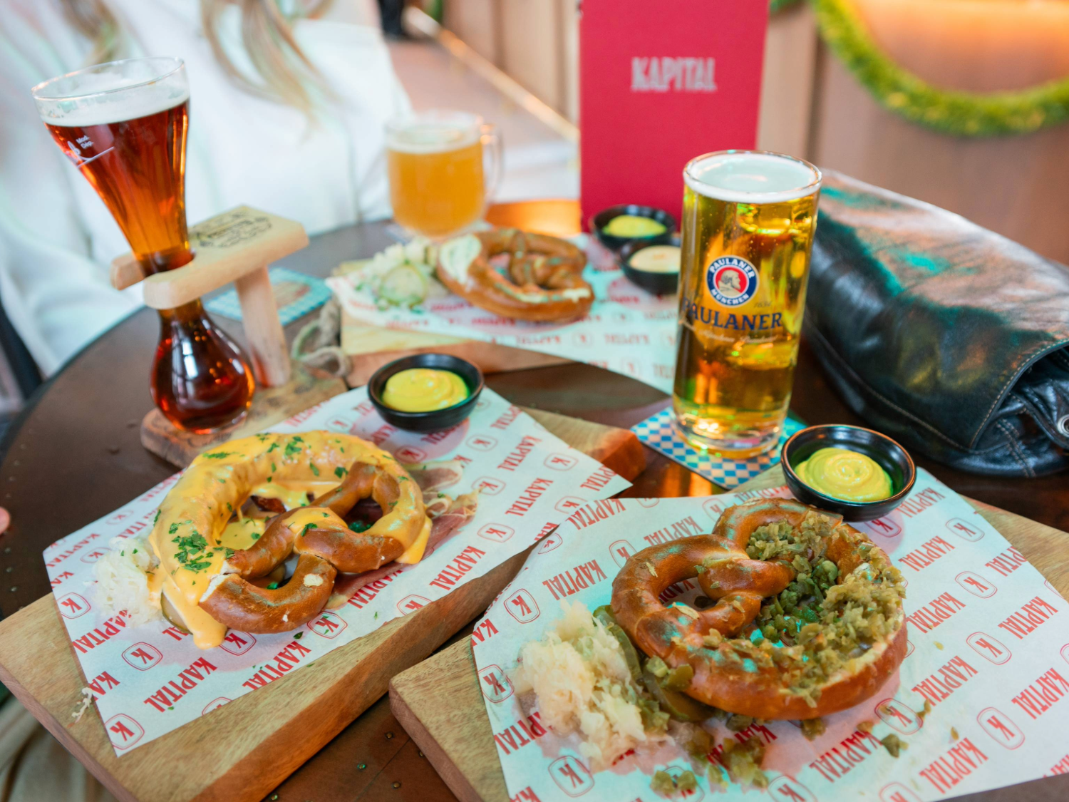 Three wooden boards on a round table display soft pretzels topped with different garnishes, served on branded paper with small pots of mustard dip. Drinks include a tall glass of dark beer, a large mug of pale beer, and a pint of Paulaner lager. A red menu with “KAPITAL” printed on it is visible in the background, along with a black handbag. The setting suggests a casual beer hall atmosphere.