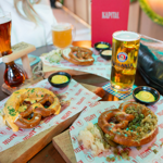 Three wooden boards on a round table display soft pretzels topped with different garnishes, served on branded paper with small pots of mustard dip. Drinks include a tall glass of dark beer, a large mug of pale beer, and a pint of Paulaner lager. A red menu with “KAPITAL” printed on it is visible in the background, along with a black handbag. The setting suggests a casual beer hall atmosphere.
