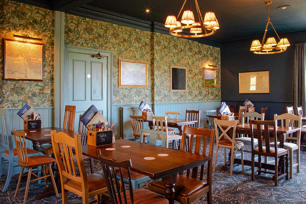 A traditional pub dining room with patterned wallpaper, wooden tables and mixed wooden chairs, lit by warm pendant lights and decorated with framed artwork.