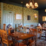 A traditional pub dining room with patterned wallpaper, wooden tables and mixed wooden chairs, lit by warm pendant lights and decorated with framed artwork.