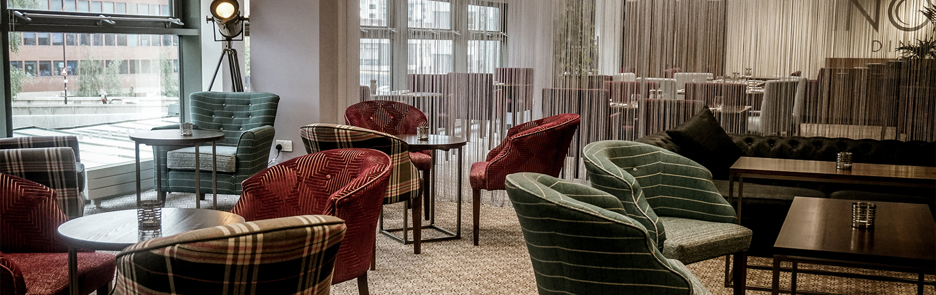 The interior of Inox Dine. Modern lounge area with patterned carpet, round tables, and upholstered chairs in various plaid and striped designs. The space features large windows providing natural light, a decorative floor lamp, and a semi-transparent curtain dividing the room. In the background, there are more tables and chairs arranged for dining, with pendant lights hanging from the ceiling.