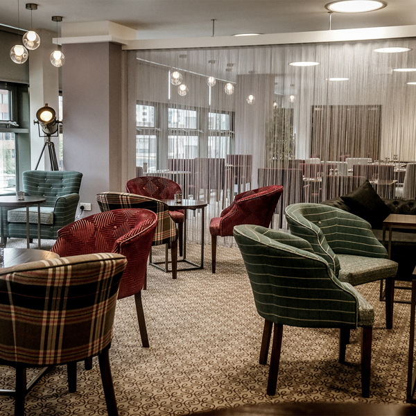 The interior of Inox Dine. Modern lounge area with patterned carpet, round tables, and upholstered chairs in various plaid and striped designs. The space features large windows providing natural light, a decorative floor lamp, and a semi-transparent curtain dividing the room. In the background, there are more tables and chairs arranged for dining, with pendant lights hanging from the ceiling.