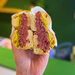A close-up of a hand holding a bagel that has been cut in half. The bagel is filled with salt beef, pickles and mustard and is wrapped in grease proof paper.
