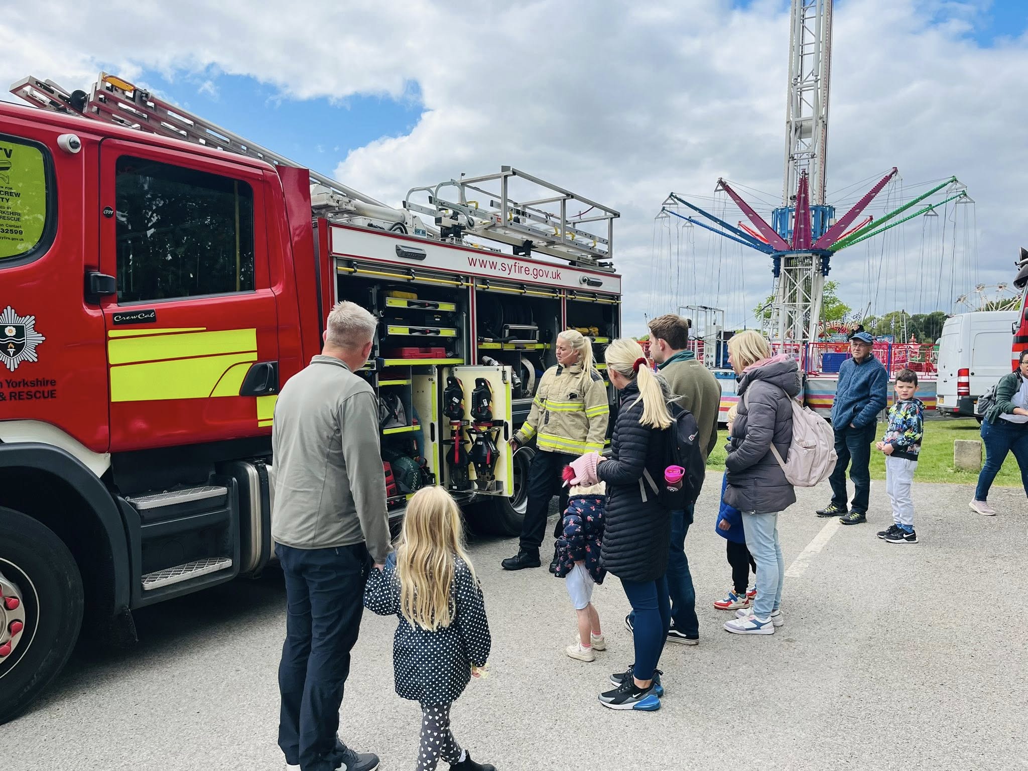 A firefighter stands beside an open fire engine showing equipment to a group of people, including adults and children, at an outdoor event. The bright red fire engine has yellow reflective stripes and ladders on top. In the background, there are fairground rides, including a tall swing ride with colourful arms, and other visitors walking around under a partly cloudy sky.