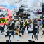 A firefighter stands beside an open fire engine showing equipment to a group of people, including adults and children, at an outdoor event. The bright red fire engine has yellow reflective stripes and ladders on top. In the background, there are fairground rides, including a tall swing ride with colourful arms, and other visitors walking around under a partly cloudy sky.