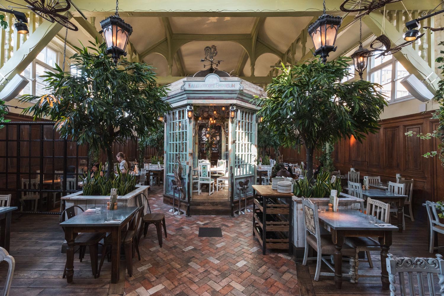 Inside The Botanist  in Sheffield.  Elegant restaurant interior with a vintage conservatory-style structure at the center, surrounded by wooden tables and chairs. The space features tall green plants, hanging lantern-style lights, and a patterned brick floor. Large windows allow natural light to fill the room, and the decor combines rustic wood panelling with ornate details for a garden-inspired atmosphere.
