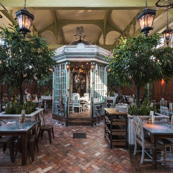 Inside The Botanist  in Sheffield.  Elegant restaurant interior with a vintage conservatory-style structure at the center, surrounded by wooden tables and chairs. The space features tall green plants, hanging lantern-style lights, and a patterned brick floor. Large windows allow natural light to fill the room, and the decor combines rustic wood panelling with ornate details for a garden-inspired atmosphere.