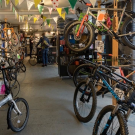 Bikes that are for sale and hire are hung on the walls and showcased on the shopfloor. A staff member is talking to a male customer in the background.