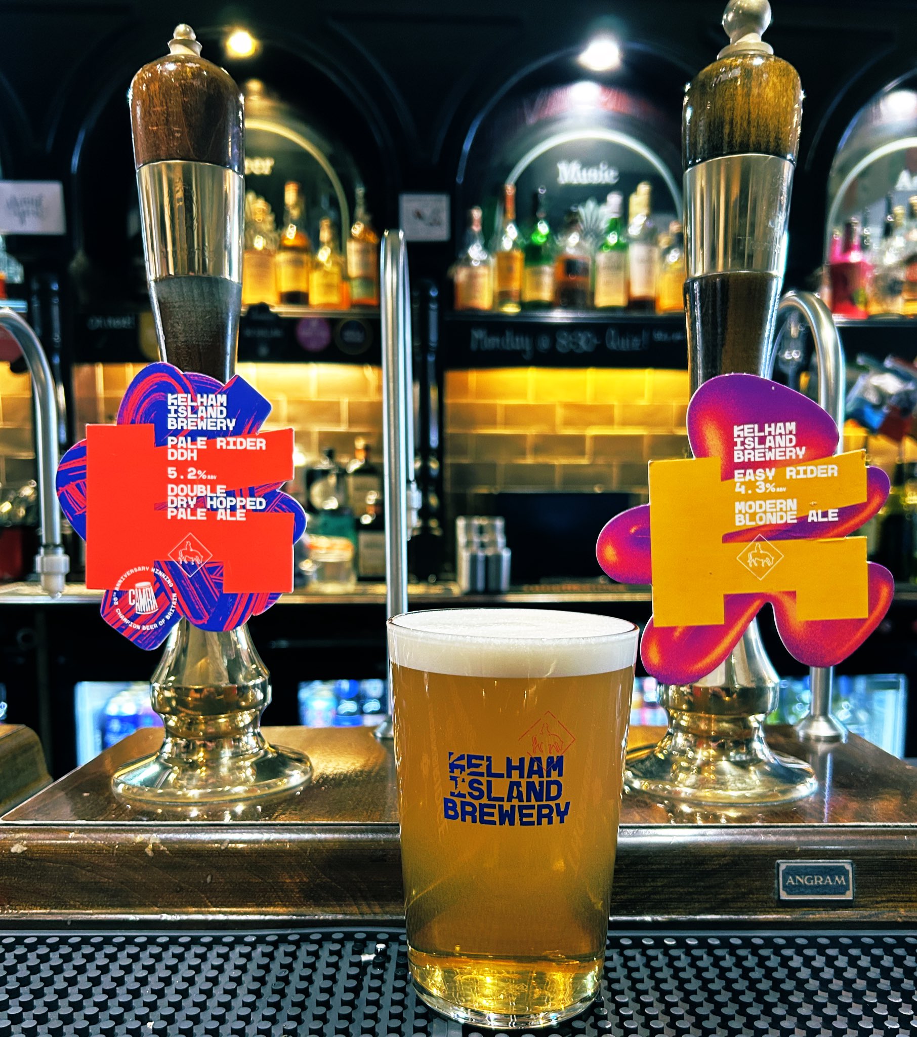 A pint of beer in a glass with the Kelham Island Brewery logo sits on a bar counter between two traditional beer pumps. The pump clips display colorful designs for two beers: one labeled “Easy Rider” Modern Blonde Ale and the other “Double Dry-Hopped Pale Ale.” The background shows shelves with liquor bottles and a warm, amber-lit backsplash.