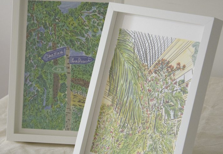 Two framed art prints displayed on a light fabric surface. The left print features a green leafy scene with a signpost pointing to “Sheffield” and “Peak District.” The right print shows a botanical illustration of tall green plants under a glass roof structure, with soft yellow and pink tones in the background.