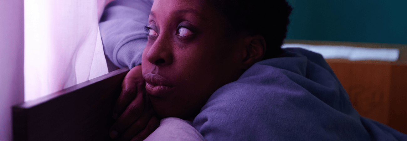 Person wearing a purple sweatshirt is leaning on a wooden bed frame near a window with sheer pink curtains. The scene is softly lit with a mix of purple and teal tones, creating a calm indoor atmosphere. The individual’s arm rests on the frame while looking toward the light coming through the curtain.