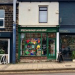 Front-facing street view of Hillsborough Bookshop situated between neighbouring buildings. The shop has a green façade with a large display window filled with brightly coloured artwork, and the sign ‘HILLSBOROUGH BOOKSHOP’ is positioned above the window and door.