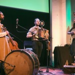 A small group of three performers stand on a stage lit with warm and green lighting. On the left, one performer holds a large upright string instrument while standing behind a large bass drum placed on the floor. In the centre, another performer plays a small accordion‑type instrument and stands in front of a microphone. On the right, a third performer plays a stringed guitar‑like instrument and stands near a microphone stand. Behind them is a table with audio equipment and balloons shaped like hearts.