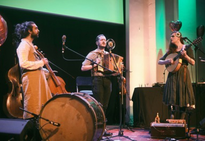 A small group of three performers stand on a stage lit with warm and green lighting. On the left, one performer holds a large upright string instrument while standing behind a large bass drum placed on the floor. In the centre, another performer plays a small accordion‑type instrument and stands in front of a microphone. On the right, a third performer plays a stringed guitar‑like instrument and stands near a microphone stand. Behind them is a table with audio equipment and balloons shaped like hearts.
