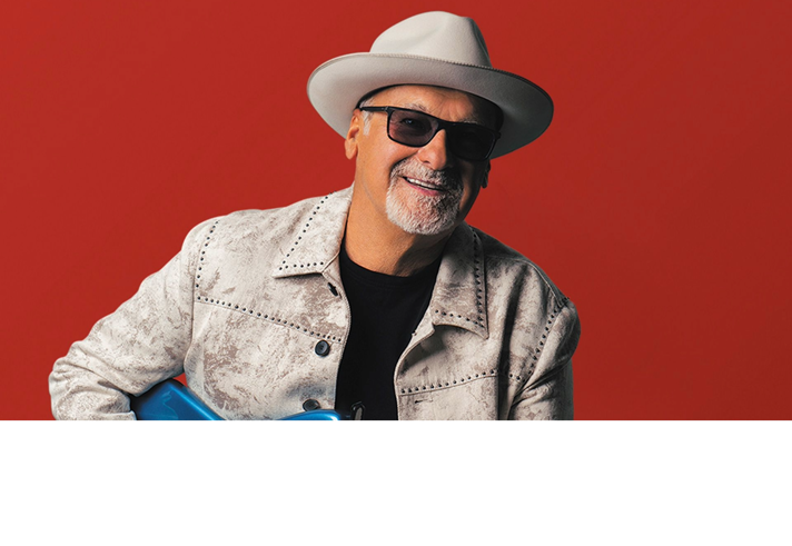 A head-shot of the artist Paul Carrack against a red backdrop.