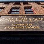 Close-up view of a large wooden sign mounted on a red-brick building. The sign reads: ‘No 20 Henry Leah & Sons Ltd, incorporating Walter Walker & Co Piercers Stampers Ltd, Cambridge Stamping Works.’ The building has tall windows and a clear blue sky above.
