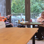 Two people seated at a table in a bright café near large glass doors with greenery visible outside. One person is holding an orange acoustic guitar, while the other is adjusting or playing a smaller string instrument. The table has mugs and papers on it, and there are chairs and wooden tables in the foreground.