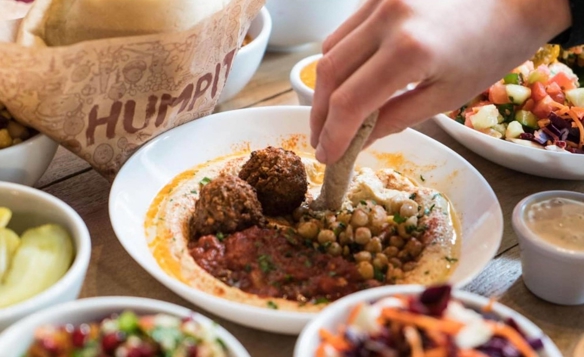 Close-up of a hand dipping pita bread into a plate of hummus topped with chickpeas, falafel, and a red sauce garnish. Surrounding the main dish are several small bowls filled with colorful salads, pickles, and toppings, along with a wrapped pita sandwich in branded paper. The table surface is wooden, and the food presentation emphasizes fresh Mediterranean flavors.